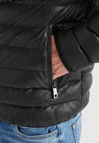 Schwarze Daunenjacke mit gestepptem Design, Reißverschluss-Tasche, glänzender Textur und einem dezenten Markenlogo am Reißverschluss.