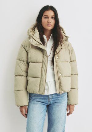 Beige gewatteerde jas met een capuchon, voorzien van een rits aan de voorkant, drukknoopsluitingen en een cropped ontwerp. Draag het over een lichtgekleurde top en spijkerbroek.
