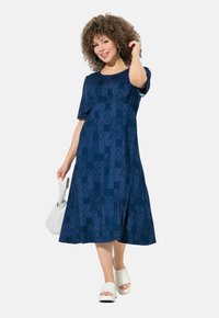 Navy jurk met korte mouwen en een geraffineerde taille, met een volledig bedrukte ontwerp. Het model houdt een witte handtas vast en draagt witte schoenen.