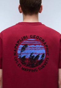 Bordeaux farbiges T-Shirt mit kurzen Ärmeln, das eine kreisförmige Grafik in Blau und Schwarz zeigt, mit einer Landschaft und dem Text: "NAPAPIJRI GEOGRAPHIC GPS ET MAPPING SURVEYS."