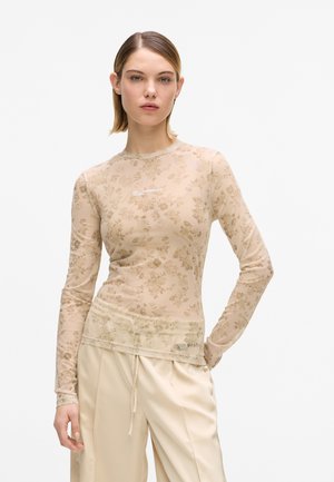 Femme portant un haut beige à manches longues avec un motif floral et le logo "Karl Lagerfeld Jeans", ainsi qu'un pantalon couleur crème sur un fond blanc.