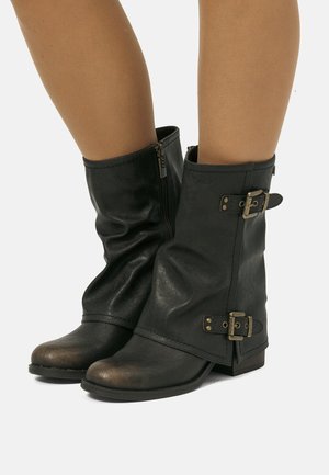 Stivali in pelle neri a metà polpaccio con fibbie e zip laterali, indossati su gambe con tonalità di pelle naturale visibili.