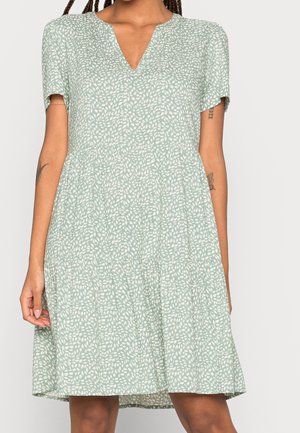 Woman wearing a short-sleeve, light green dress with a white leaf pattern and a V-neckline, showing arm tattoos and relaxed fit.