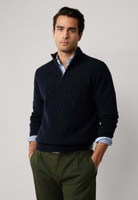Maglione navy a treccia con colletto a mezza zip, indossato sopra una camicia azzurro chiaro. Abbinato a pantaloni verde oliva, che offrono una finitura texurizzata.