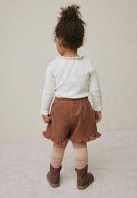 Vuggestuebarn med krøllet hår iført en hvid langærmet bluse, bølgede brune shorts, stribede strømpebukser og brune støvler, der står foran en ensfarvet baggrund.