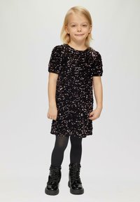 Schwarzes Paillettenkleid mit kurzen Puffärmeln, strukturierten Oberflächen, kombiniert mit schwarzen Leggings und glänzenden Kampfstiefeln, vor einem hellen Hintergrund stehend.