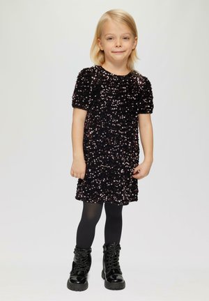 Schwarzes Paillettenkleid mit kurzen Puffärmeln, strukturierten Oberflächen, kombiniert mit schwarzen Leggings und glänzenden Kampfstiefeln, vor einem hellen Hintergrund stehend.