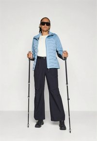 Osoba v slnečných okuliaroch, modrej bunde, bielom tričku a čiernych nohaviciach, ktorá drží dve turistické palice a stojí proti jednému svetlému pozadiu.