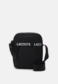Borsa a tracolla nera in tessuto con chiusura zip, tracolla regolabile e il logo bianco "LACOSTE" ben visibile sul davanti.