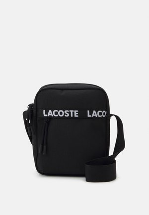 Schwarze Stoff-Crossbody-Tasche mit Reißverschluss, verstellbarem Tragegurt und dem auffälligen weißen "LACOSTE"-Logo auf der Vorderseite.