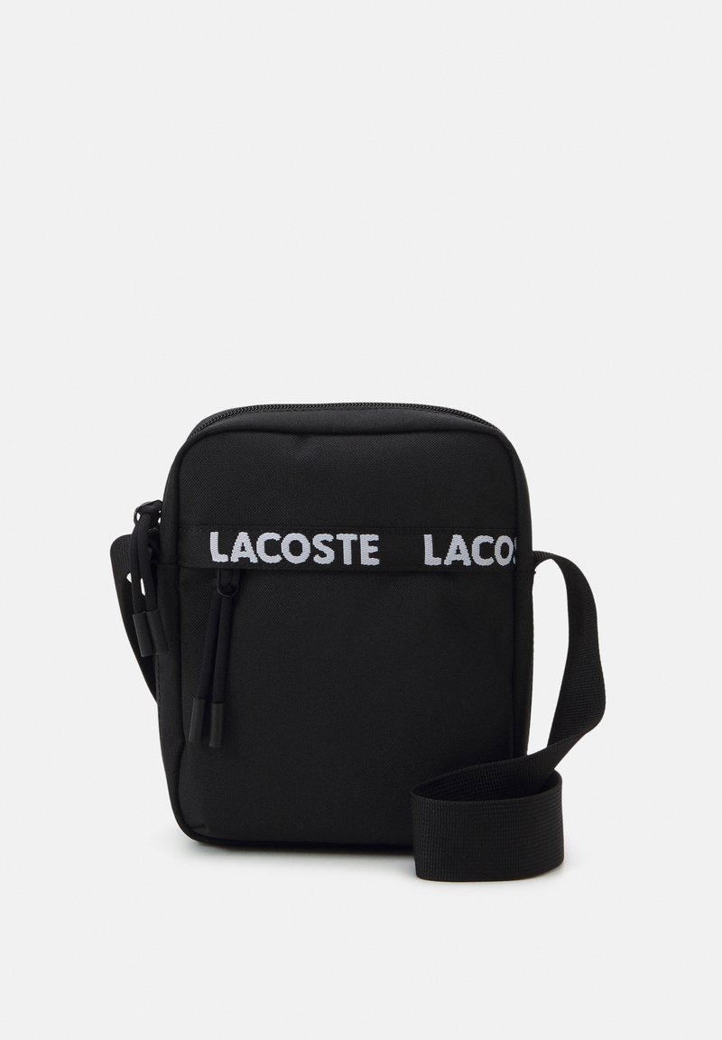 Sac bandoulière noir en tissu avec fermeture éclair, sangle réglable et logo "LACOSTE" blanc bien visible sur le devant.