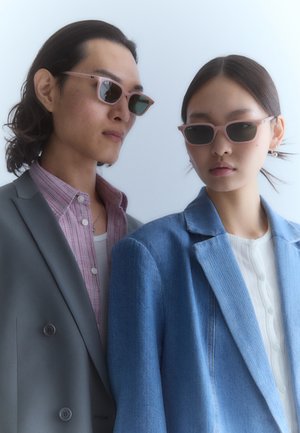 Šviesiai rožinės rėmeliais akiniai nuo saulės su tamsiais objektyvais, kuriuos dėvi du asmenys. Vienas vilki pilką kostiumą, o kitas - džinsinę striukę ant baltos marškinės.