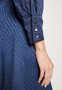 Marinblå klänning med en texturerad tyg med sexkantigt mönster. Ärmen har guldknappar som dekoration vid manschetten.