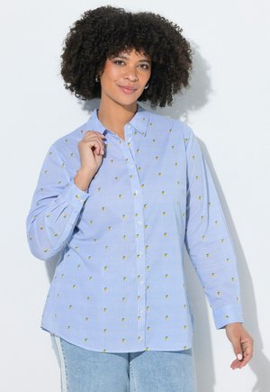 Kvinde med krøllet hår iført en blå stribet knap-op-skjorte med små gule og grønne mønstre, poserende mod en hvid baggrund.