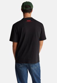 Camiseta negra de algodón con mangas cortas, con texto rojo en la parte posterior. Se lleva con vaqueros azules, vista desde atrás.