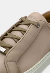 Μπεζ δερμάτινο αθλητικό παπούτσι με στρογγυλή μύτη, κορδόνια στον ίδιο τόνο και ασημένιες τρύπες για τα κορδόνια. Διαθέτει υφή στην επιφάνεια και διακριτική λεπτομέρεια με λογότυπο στο πλάι.