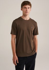 Hnědé bavlněné tričko s kulatým výstřihem a krátkými rukávy; má malé vyšívané logo na levé straně hrudi.