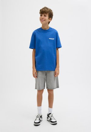 Ragazzo in piedi e sorridente, indossa una maglietta blu, pantaloncini grigi, calzini bianchi e sneakers bianche e nere su uno sfondo bianco semplice.