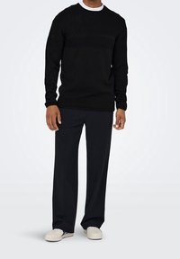 Schwarzer Strickpullover mit einem strukturierten Muster, Rundhalsausschnitt und langen Ärmeln, kombiniert mit lockeren schwarzen Hosen und weißen Sneakers.