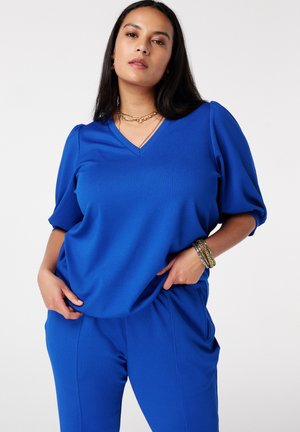 Vrouw poseert met handen in de zakken, draagt een koningsblauw V-hals topje met pofmouwen en bijpassende broek tegen een effen achtergrond.