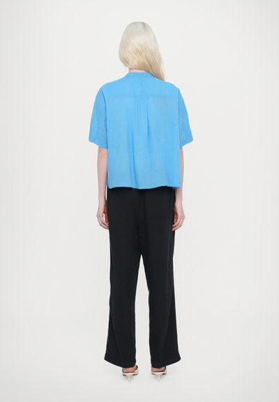 Camicia azzurra chiara a maniche corte con una vestibilità ampia, dettaglio con yoke posteriore, abbinata a pantaloni neri a gamba larga e scarpe con tacco di colore chiaro.