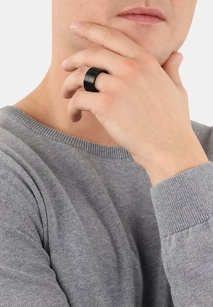 Anello in silicone nero con multiple scanalature orizzontali, indossato su un dito. Chi lo indossa ha un maglione grigio chiaro. Inquadratura ravvicinata della mano e dell'anello.