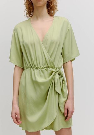 Robe d'un vert clair, style portefeuille, avec un col en V et des manches courtes. Présente une taille froncée et un détail de nœud sur le côté gauche. Tissu doux.