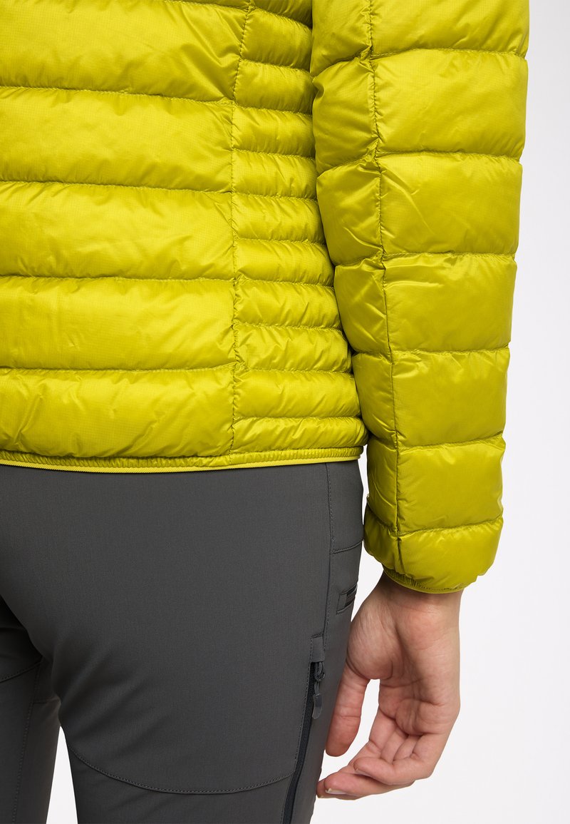 Limettengrüner, gestepptes Pufferjacke mit ausgeprägten Abschnitten; kombiniert mit dunkelgrauen Hosen mit einer Reißverschlusstasche an der Seite.