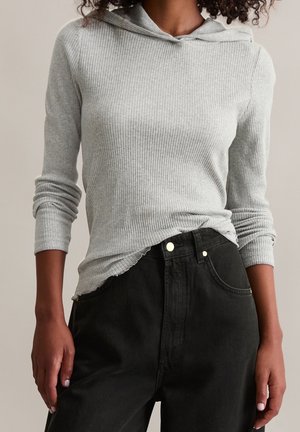 Femme portant un haut à capuche côtelé gris clair à manches longues, rentré dans un jean noir taille haute avec des boutons métalliques.