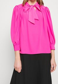 Kirkas vaaleanpunainen paita, jossa on solmukaulus, puffihihat ja joustavat hihansuut. Sileä kangas ja löysä istuvuus, yhdistettynä mustiin hameisiin.