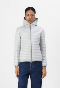 Hellgraue, gepolsterte Jacke mit hohem Kragen, Reißverschluss und Kapuze. Vertikale Streifen und glatte Textur. Getragen mit blauen Jeans.