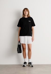 Kvinne som står mot en hvit vegg iført en svart oversized t-skjorte, hvit plisserte skjørt, hvite sokker, svarte joggesko, og holder en svart håndveske.