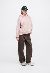 Rózsaszín kapucnis pulóver elülső zsebbel, széles lábú barna nadrággal párosítva. A fekete sapka és a szürke sportcipő teszi teljessé az öltözetet.