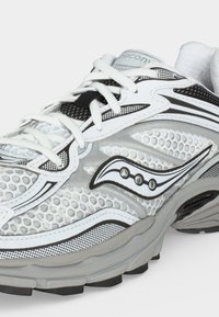 Λευκό και γκρι αθλητικό παπούτσι Saucony με άνω μέρος από δίχτυ, ασπρόμαυρο λογότυπο, λευκά κορδόνια και μαξιλαρωτή σόλα.