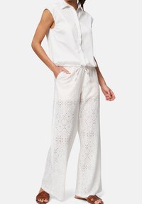 Chemise blanche sans manches avec fermeture à boutons sur le devant, associée à un pantalon blanc à jambes larges avec motif en dentelle, et des sandales marron, doté de poches et d'une taille à cordon.