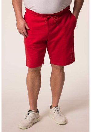 Adulto de pie vistiendo pantalones cortos rojos brillantes con cordón, zapatillas deportivas blancas y una camisa clara metida por dentro, sobre un fondo liso.