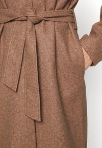 Brun filthatte med båndlukning i taljen, med en glat tekstur og foldet reversdesign. Inklusiv sidelommer.