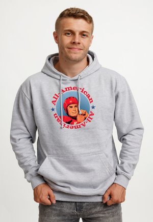Mężczyzna w jasnoszarej bluzie z kapturem z grafiką piłkarza w czerwonym hełmie oraz napisem "All-American" wokół obrazka w kolorze czerwonym.