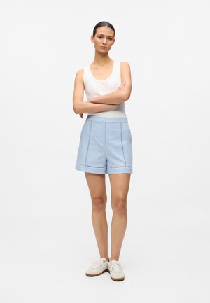 Svetlomodré šortky na mieru s hladkým povrchom, bočnými vreckami a dekoratívnym prešitím pozdĺž lemu. Kombinované s bielym tielkom.