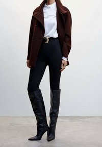 Manteau en laine bordeaux porté sur un haut blanc côtelé, associé à des leggings noirs taille haute et des bottes stiletto en cuir noir montant jusqu'aux genoux.