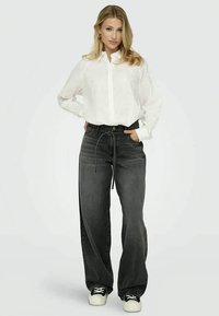 Camisa branca de manga longa com textura subtil, combinada com jeans cinzentos escuros de perna larga e ténis pretos.