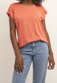 Korálové tričko s krátkým rukávem a kulatým výstřihem, vyrobené z měkkého materiálu. Kombinováno s džíny světle modré barvy s rovnými nohavicemi. Hladká textura.