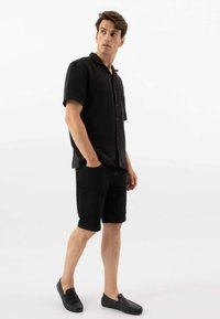 Zwart kortmouwen overhemd met knoopjes en zak, gecombineerd met zwarte shorts en zwarte instappers, gepresenteerd tegen een neutrale achtergrond.