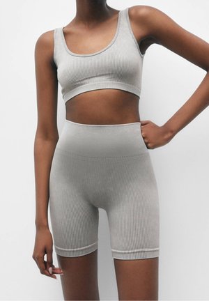 Vrouw die een bijpassende lichtgrijze geribbelde cropped top en een hoge taille biker shorts draagt tegen een effen witte achtergrond.