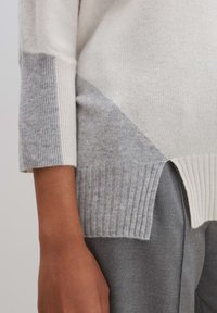 Leichter Strickpullover in Creme und Hellgrau, mit einem diagonalen Farbblockdesign, gerippten Bündchen und seitlichen Schlitzen für zusätzliche Details.