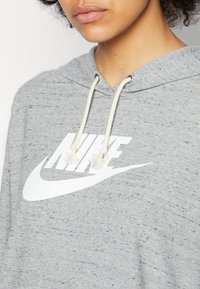 Felpa grigia con cappuccio e logo Nike bianco. Presenta un cappuccio con cordino e ponpon, una texture morbida e una vestibilità comoda.