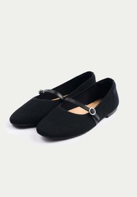Svarta tygballerina med ribbad textur, spetsig tå och en tunn svart rem med en silverfärgad cirkulär detalj.