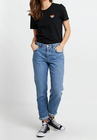 Kvinde iført sort t-shirt med en lille hjertepåsyning, lyseblå optrillede jeans og sorte høje sneakers står med hænderne i lommerne.