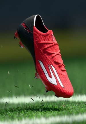 Scarpa da calcio rossa e nera con logo bianco, sospesa in aria sopra un campo d'erba con zolle sollevate e linea di confine bianca visibile.