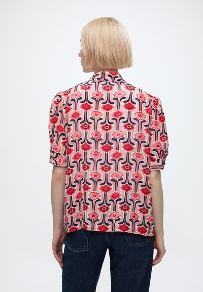 Femme aux cheveux blonds courts portant un chemisier à manches courtes orné de fleurs rouges et roses, associé à un jean bleu foncé.
